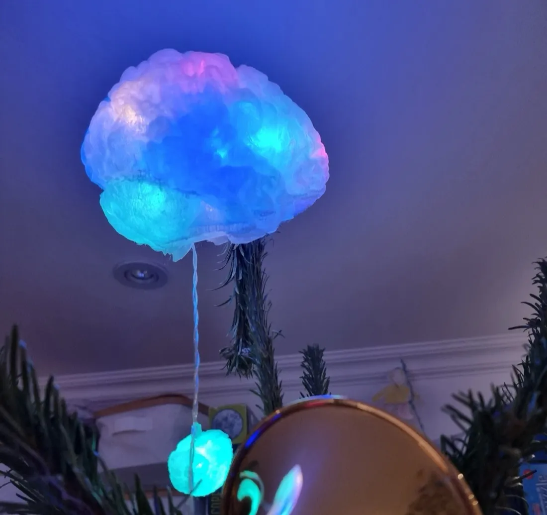 A 3D-printed brain lit from inside, glowing blue, cyan and pink against a ceiling at the top of a Christmas tree, with a smaller lit print hanging below it and a trumpet bell catching the light.