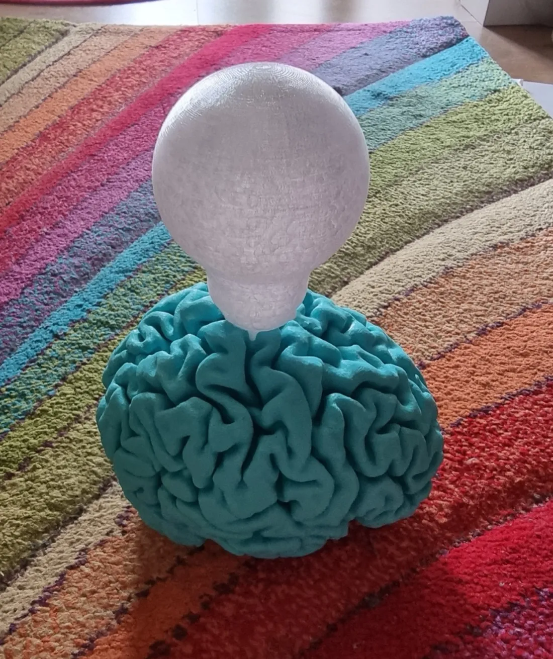 A teal 3D-printed brain with its folds clearly resolved, sitting on a striped rainbow rug, with a smooth white printed light bulb balanced on top of it.