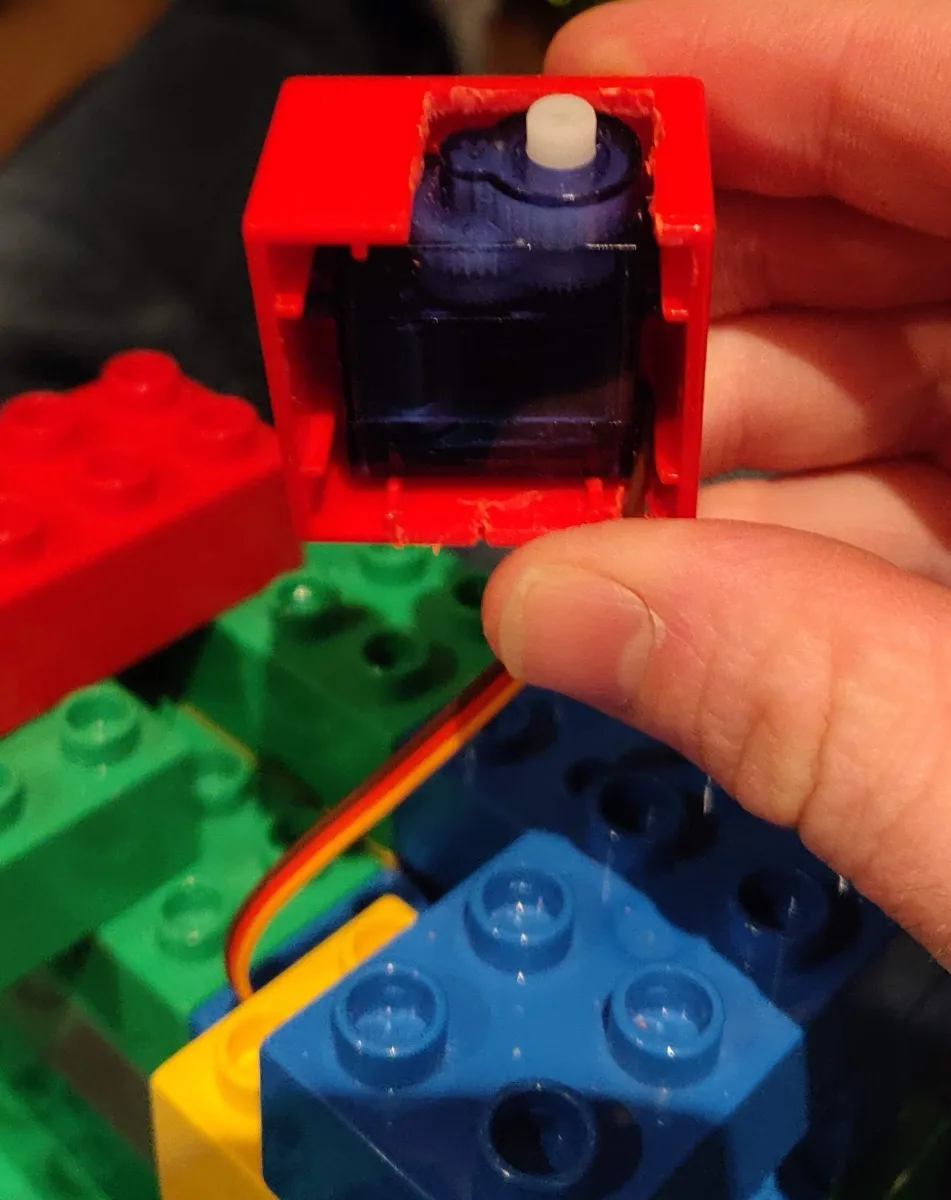 A red Duplo brick with one face cut away and a micro servo seated inside it, held above a part-built Duplo tower.