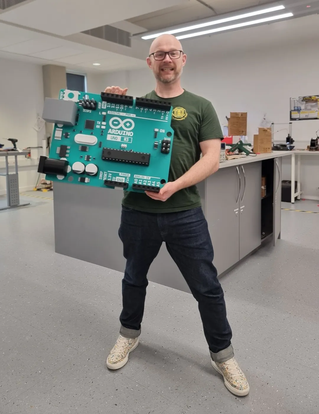 Andy standing in a fabrication lab holding a 3D-printed Arduino Uno R3 roughly the width of his shoulders, with the headers, silkscreen labels and microcontroller all reproduced.