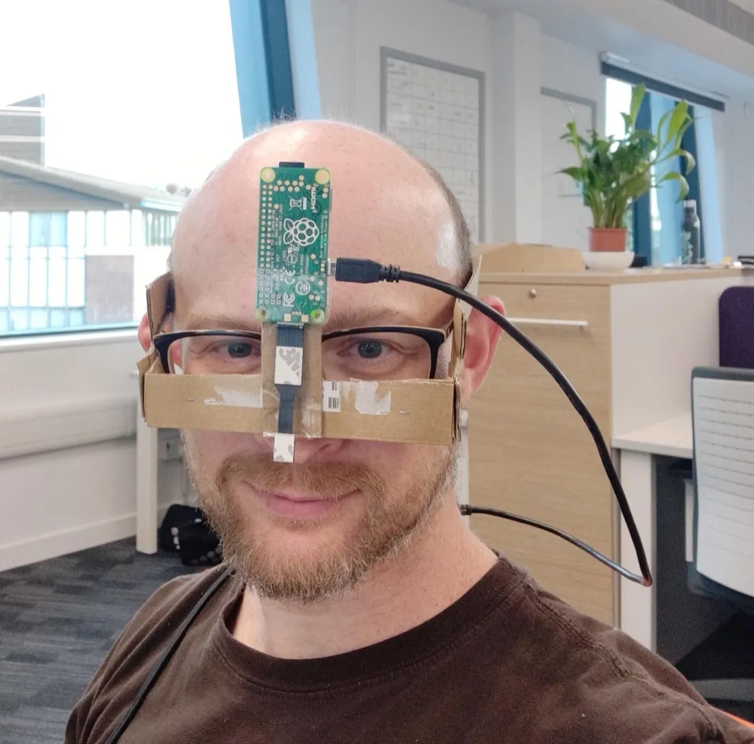 The early prototype worn in an office: a Raspberry Pi Zero W and a camera ribbon cable taped to a cardboard frame fixed across a pair of glasses.