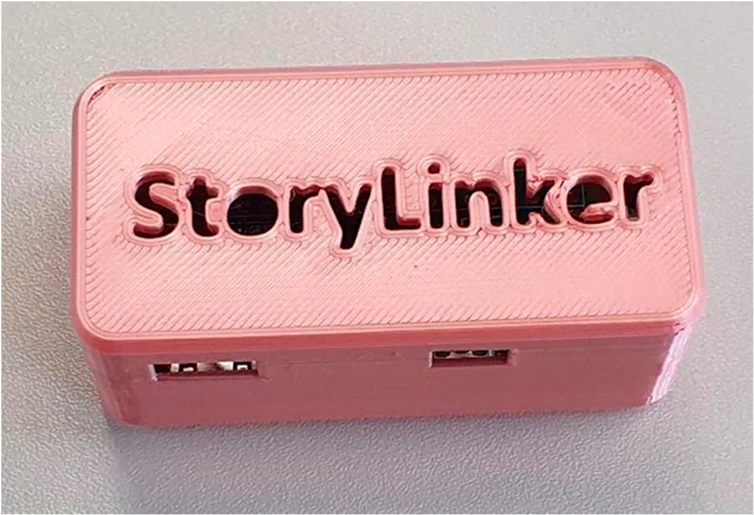 The StoryLinker dongle: a small pink 3D-printed enclosure with StoryLinker embossed on the lid, two USB ports showing through cut-outs in the side.
